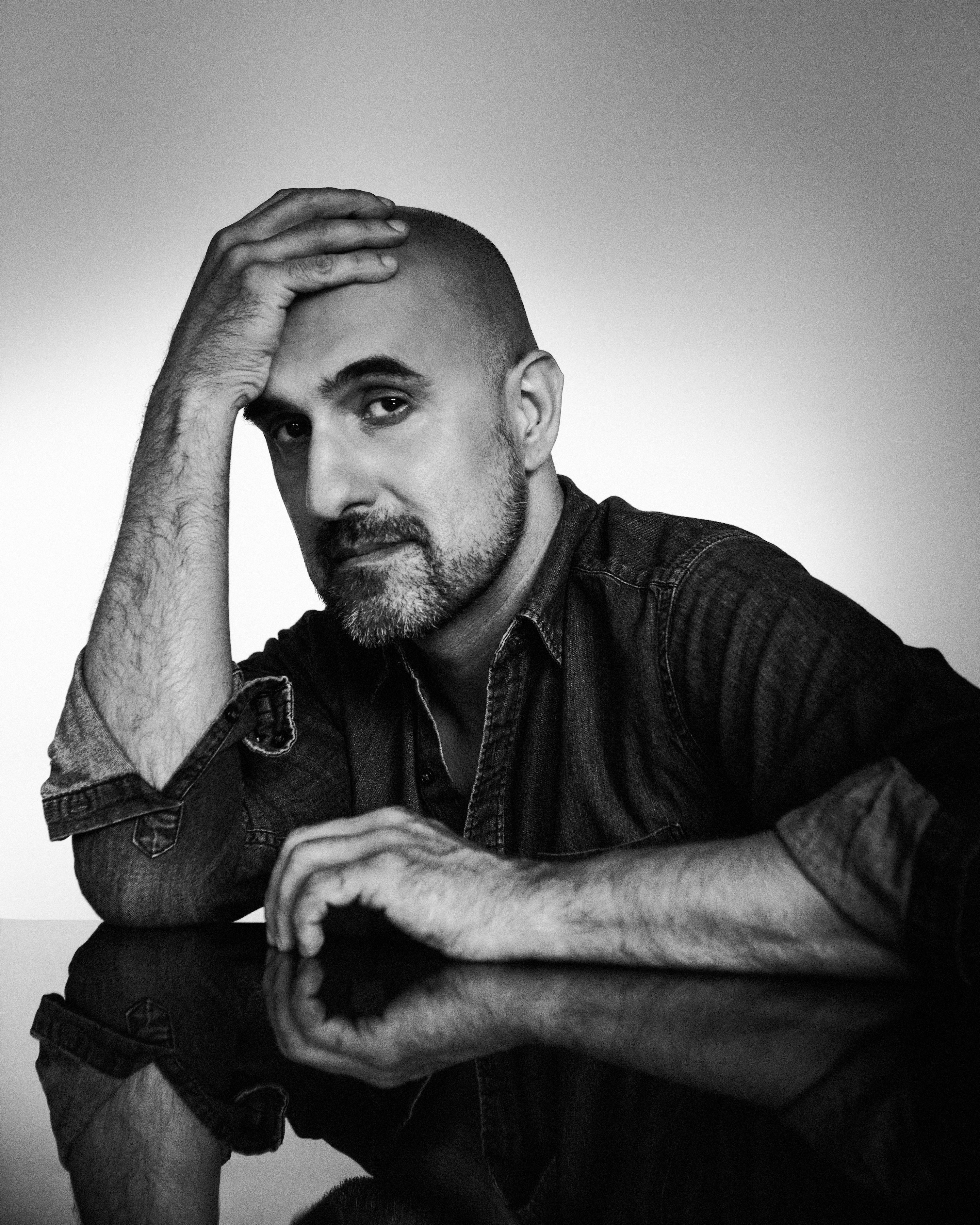 A black and white photo. Novelist Hari Kunzru sits, wearing a dark shirt, one arm flat on a reflective table top and his other elbow resting on the table with his head resting on his hand.