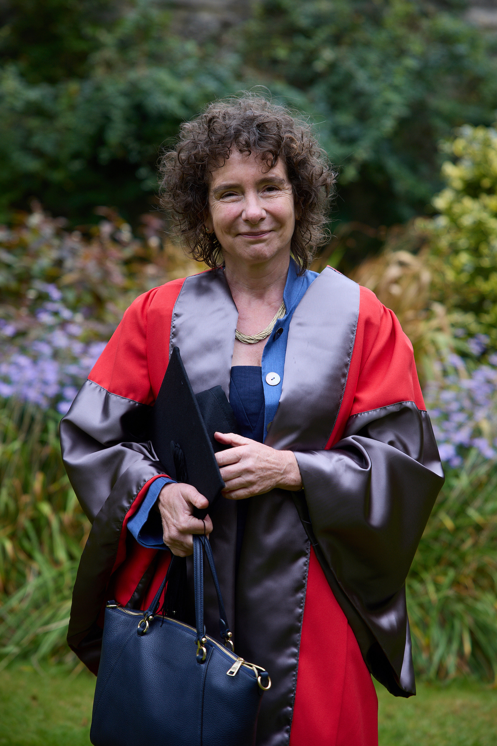 Jeanette Winterson stands in a garden wearing the bright red and grey robes for Encaenia, having received an honorary degree in 2021