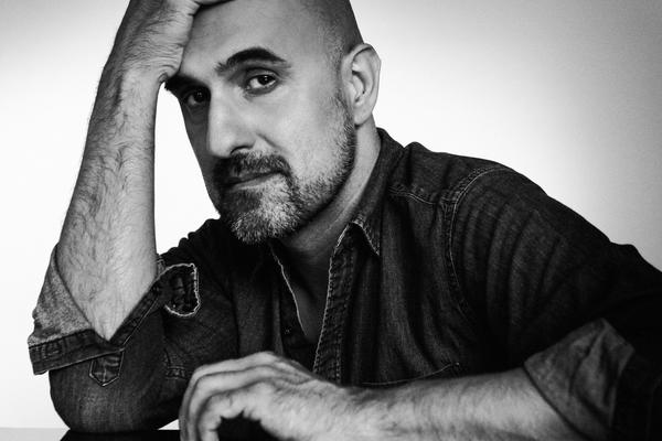 A black and white photo. Novelist Hari Kunzru sits, wearing a dark shirt, one arm flat on a reflective table top and his other elbow resting on the table with his head resting on his hand.