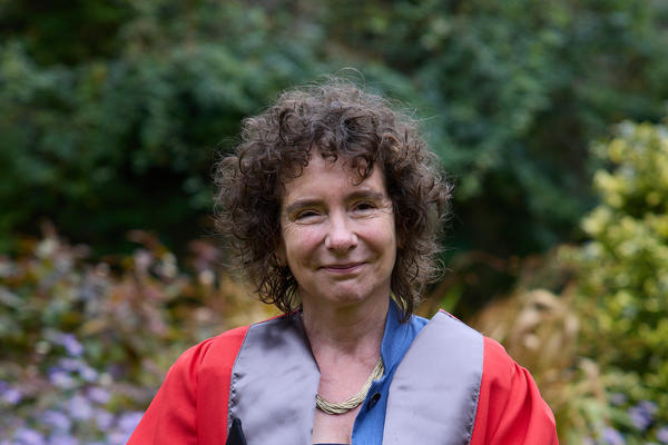 Jeanette Winterson stands in a garden wearing the bright red and grey robes for Encaenia, having received an honorary degree in 2021