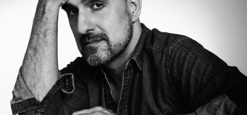 A black and white photo. Novelist Hari Kunzru sits, wearing a dark shirt, one arm flat on a reflective table top and his other elbow resting on the table with his head resting on his hand.