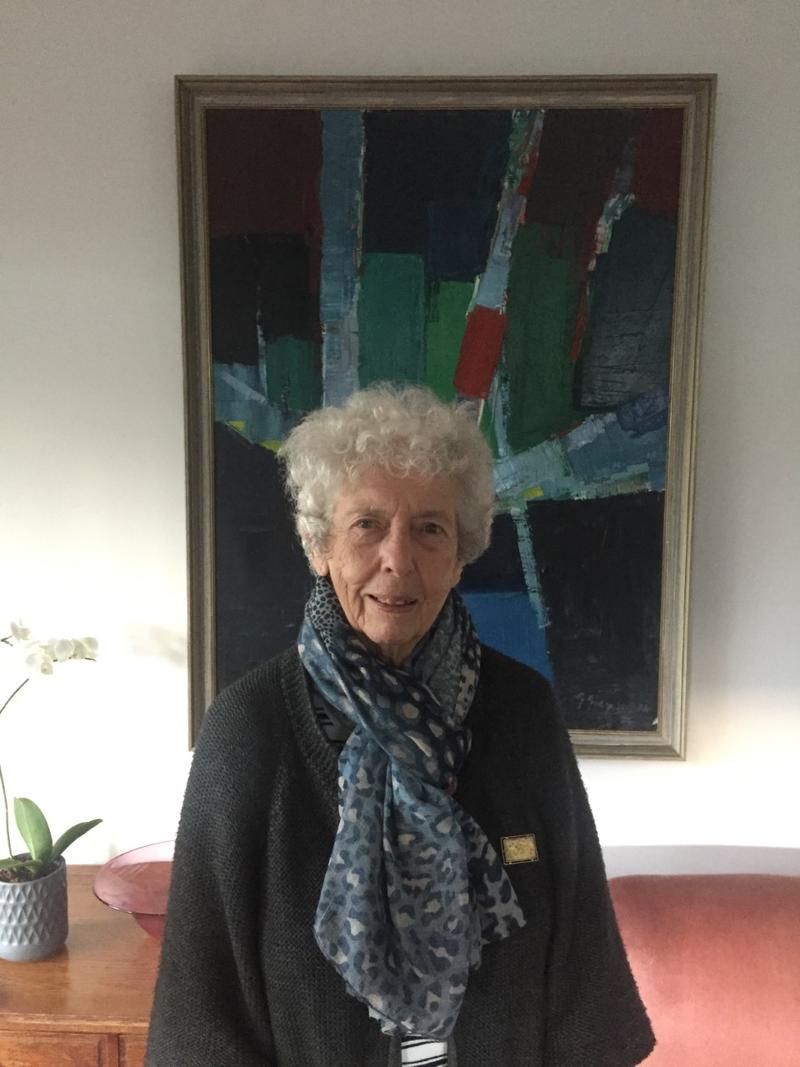 Carol Bolton, wearing a dark jacket and blue patterned scarf, stands in front of an abstract painting of green, red and blue