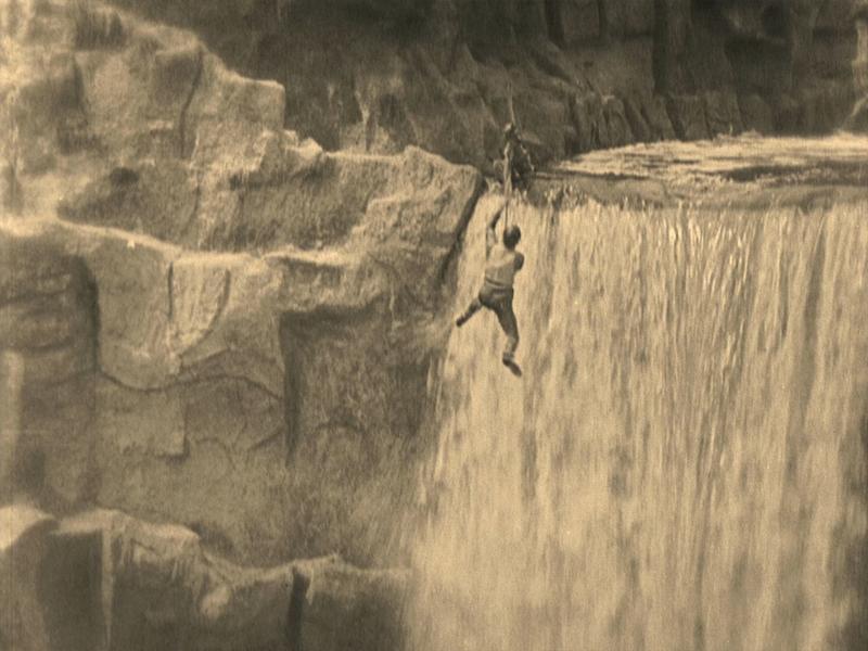 A black and white film still of Buster Keaton dangling from a rope in front of a waterfall
